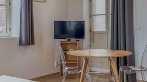 Una habitación con una televisión sobre un mueble de madera, una mesa redonda de madera con sillas, un reloj en la pared y una puerta con paneles de vidrio esmerilado.