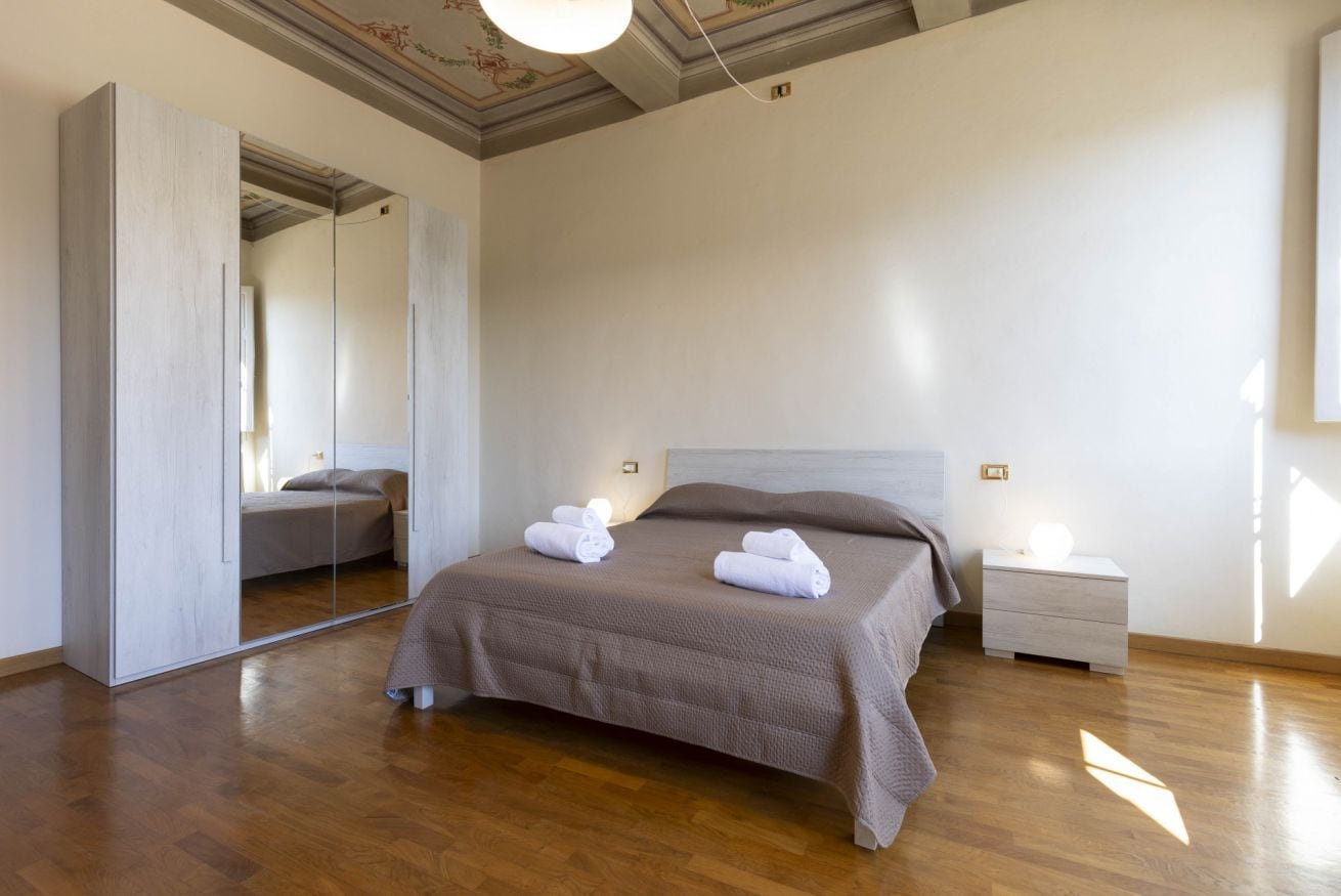 Een slaapkamer met een groot bed, nachtkastje, spiegel en een plafondlamp.
