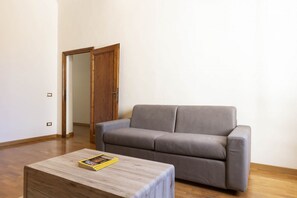Een woonkamer met een grijze bank, een houten salontafel en een houten deur die naar een andere kamer leidt.