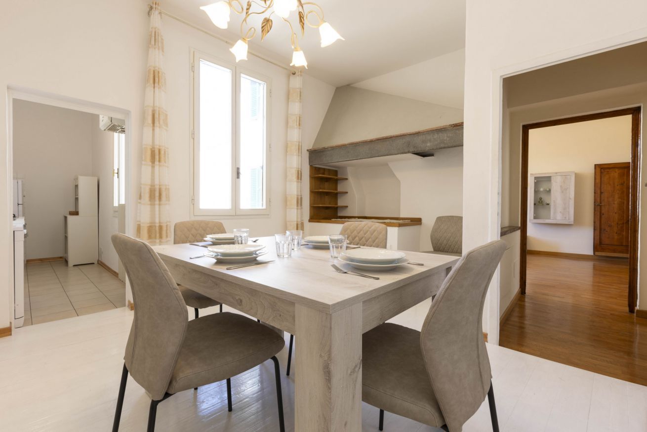 Een eetruimte met een houten tafel, gedekt voor vier personen, omringd door stoelen. Achtergrond is een keuken met een inbouwkast.