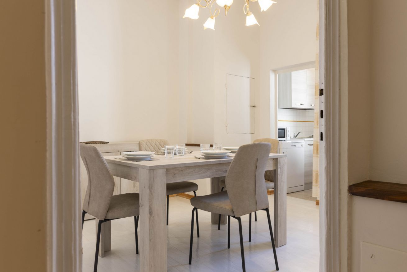 Een eetgedeelte met een houten tafel en vier stoelen, een keuken op de achtergrond, en een witte inrichting.