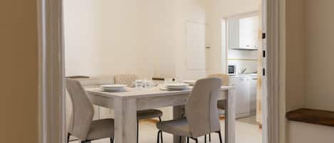 Un comedor con una mesa de madera y cuatro sillas, una cocina al fondo, y un interior blanco.