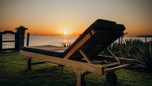 Una silla de lounge de madera con cojín, ubicada en un área verde con vista al atardecer sobre el agua.