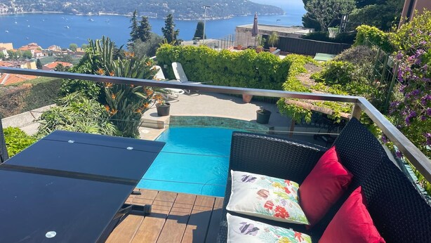 Uma varanda com piscina, um sofá de vime com almofadas e vista para o mar e as colinas.
