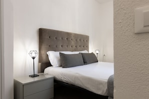 A modern bedroom with a tufted headboard, a bed with white and gray bedding, two bedside tables with lamps, and a small drawer unit. - Santo Spirito Living (Firenze)
