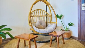 Een hangstoel van riet met een kussen, twee houten bijzettafeltjes en een bloempot.