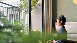 Iemand zit bij een raam met uitzicht op een balkon en bloempotten.