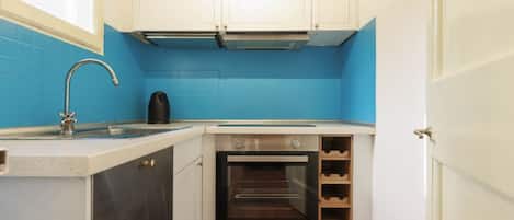 Cocina compacta con armarios blancos, salpicadero azul, pileta de acero inoxidable, tetera negra y horno integrado.