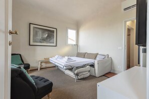 Une chambre d’hôtel compacte comprenant un lit pliable, un petit bureau, une chaise, un tableau encadré au mur et une fenêtre laissant entrer la lumière naturelle.