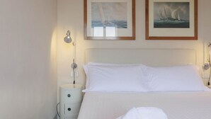 Ein ordentlich gemachtes Bett mit weißen Leinen, eine Nachttischlampe und zwei gerahmte Bilder an der Wand.