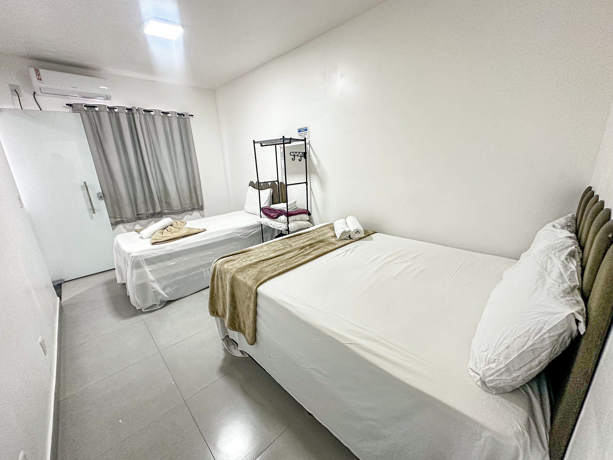 Quarto de hotel com duas camas de solteiro, guarda-roupa, espelho e janela com cortinas.