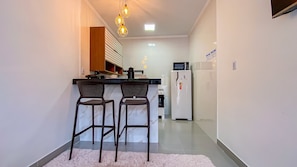 A compact kitchen with a refrigerator, microwave, and a coffee maker. There are two bar stools and a wooden cabinet above the counter. - Condomíno American Praia  (Porto Seguro)