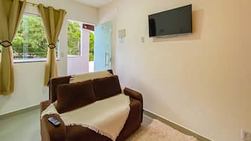 Sala de estar com um sofá marrom, um controle remoto sobre ele, uma TV fixada na parede e uma porta dando para o exterior.