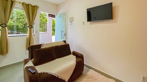 A living room with a brown sofa, a remote control on it, a TV mounted on the wall, and a door leading outside. - Condomíno American Praia  (Porto Seguro)