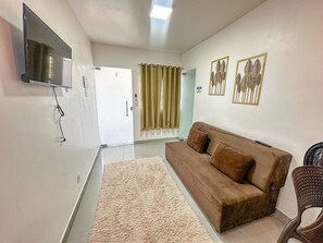 A living room with a brown sofa, a flat-screen TV mounted on the wall, a beige rug, and two framed wall art pieces. - Condomíno American Praia  (Porto Seguro)