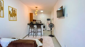 Ein kompakter Wohnbereich mit einer Küchenzeile, einem Essbereich mit Barhocker, einem an der Wand befestigten Fernseher und einem Standventilator.