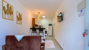 Ein modernes Wohnzimmer mit einem braunen Sofa, einem Essbereich mit Tisch und Stühlen, einer Küche inklusive Kühlschrank und Mikrowelle sowie einem an der Wand montierten Fernseher.