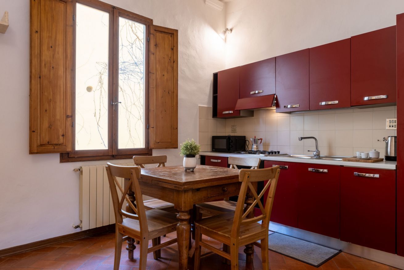 Une cuisine avec des meubles de rangement en bois, une table à manger avec des chaises, une plante en pot et une fenêtre avec des volets en bois.