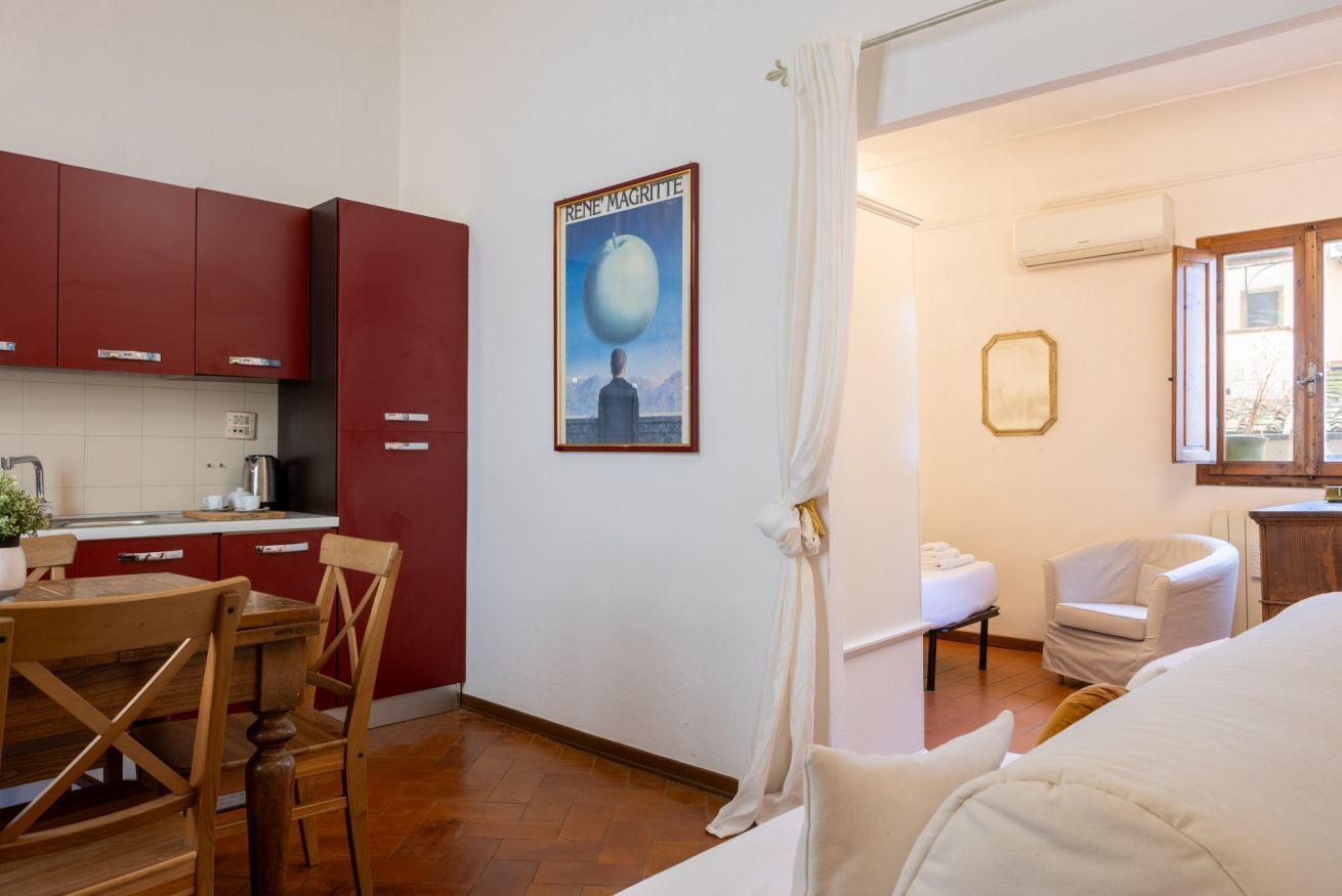 Una stanza con un tavolo da pranzo in legno, pensili rossi, un quadro, un letto, una sedia e una finestra.
