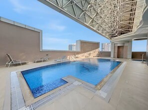 Une piscine intérieure dotée d’une structure de plafond aux formes géométriques, un espace aménagé au bord de la piscine avec des chaises longues, et une vue sur les gratte-ciel en arrière-plan.