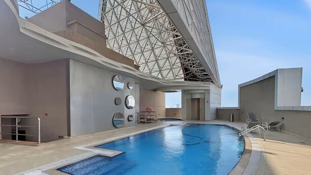 An indoor swimming pool with a modern architectural design, including a geometric glass ceiling and a poolside area with lounge chairs. - Vogue | Siraj Tower Arjan near Miracle Garden (Dubai)