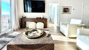 Ett modernt vardagsrum med en stor TV, en vit soffa, ett soffbord med tidningar och utsikt över naturen genom ett stort fönster.