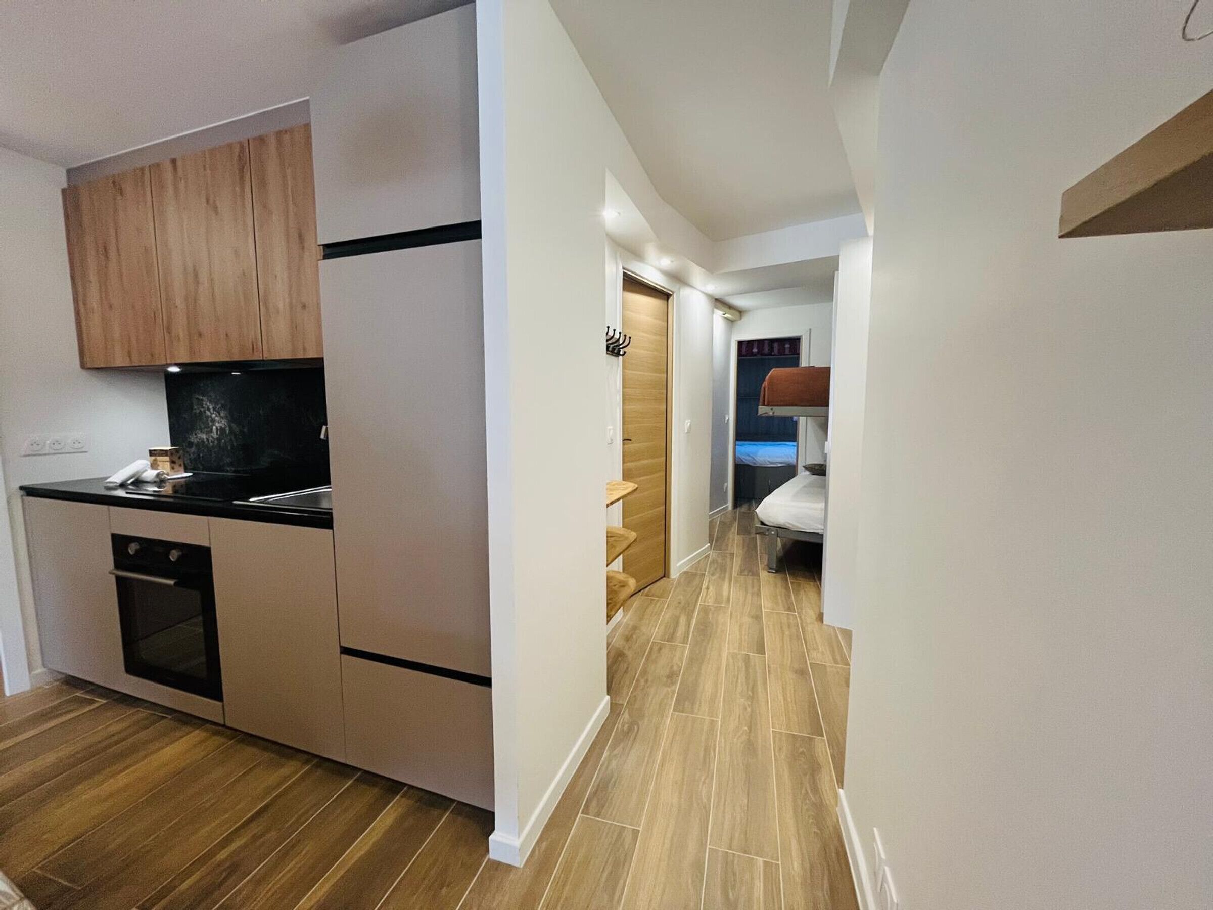 A compact kitchen with modern appliances, a wooden cabinet, and a small oven. A narrow hallway leads to a bedroom with a bunk bed.