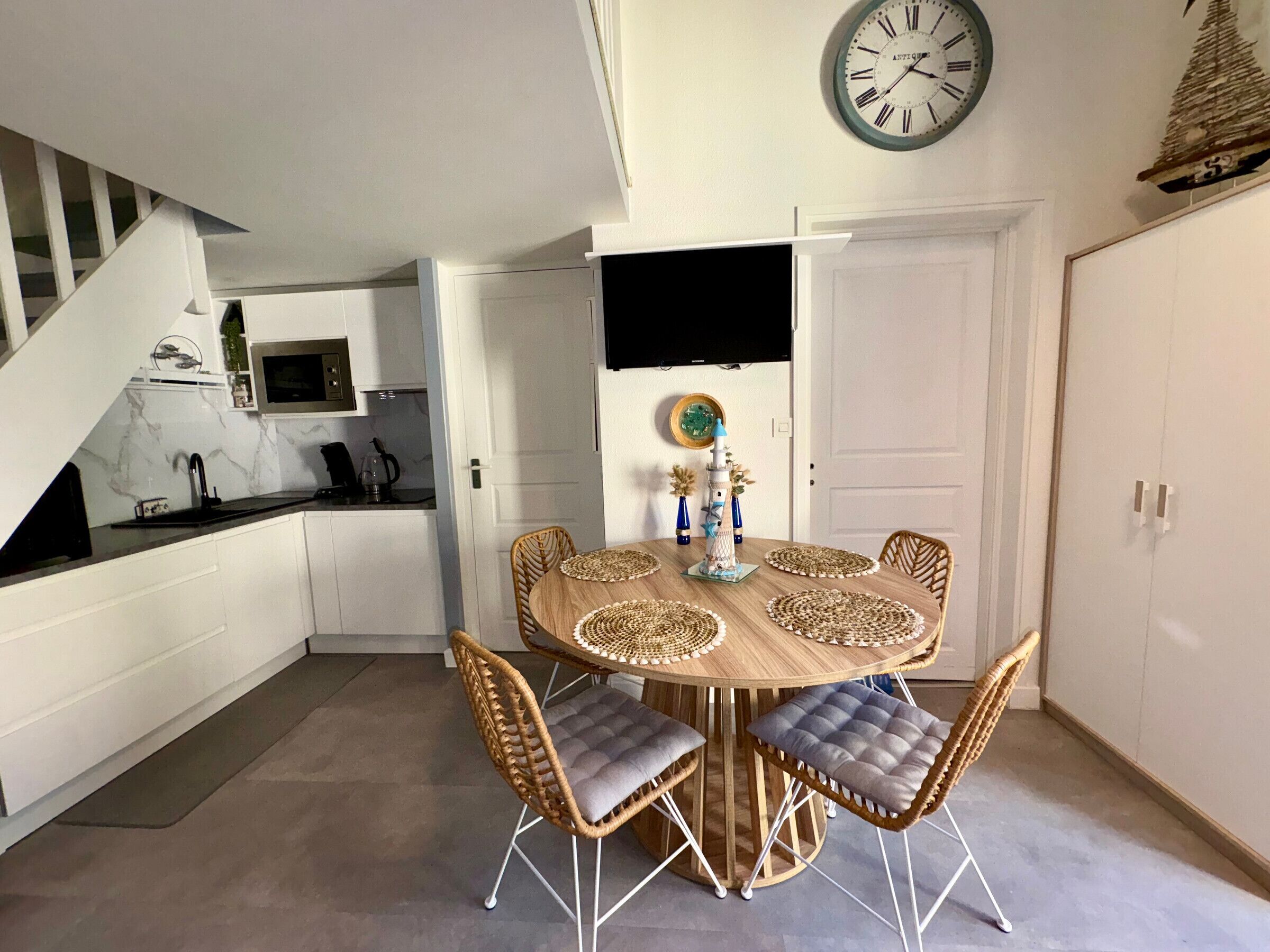 Una cocina moderna con armarios blancos, encimera negra y un comedor con una mesa redonda de madera y sillas de mimbre.