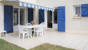Un patio con mesa y sillas blancas, contraventanas azules y un toldo a rayas.