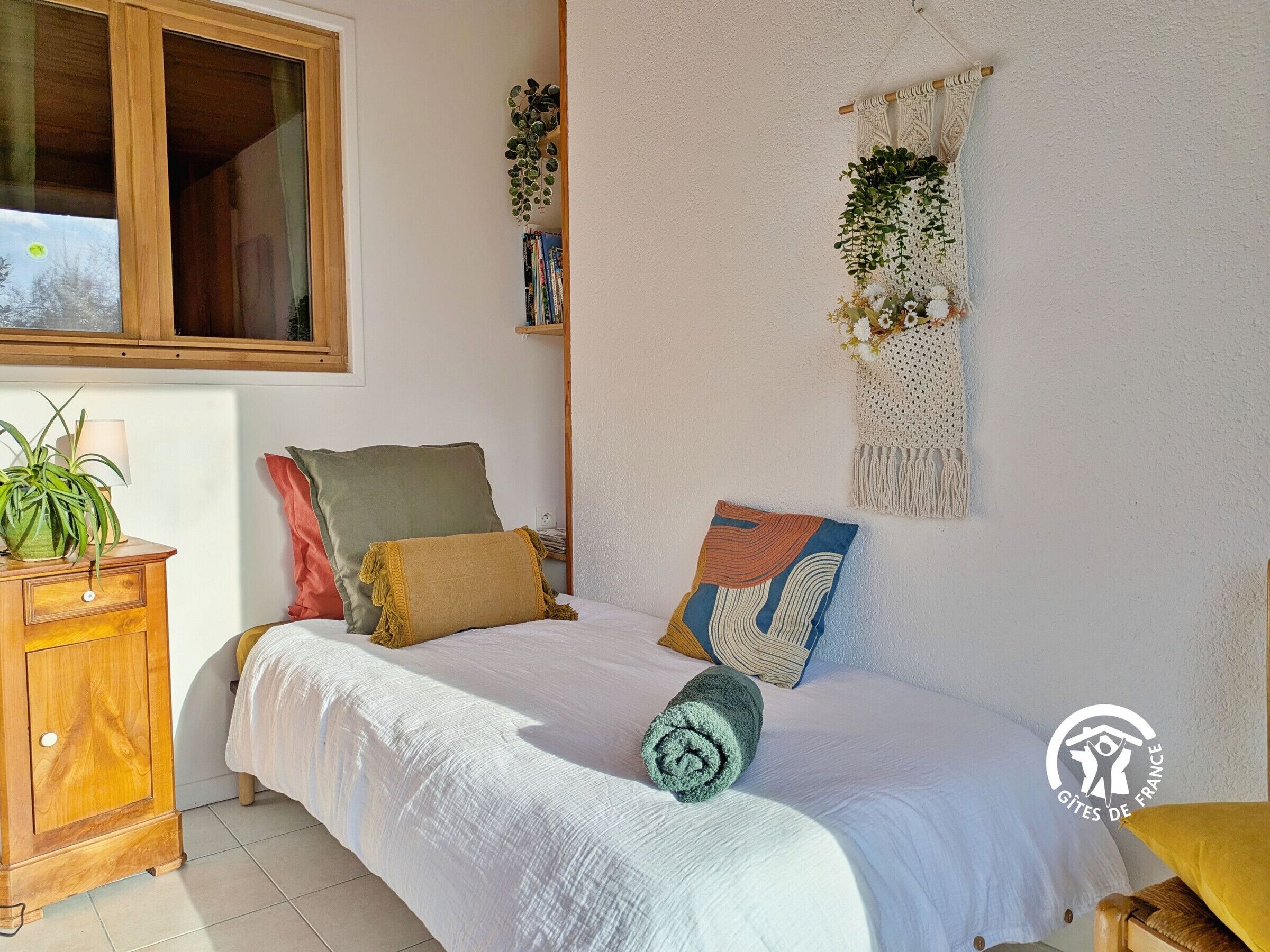 A bedroom with a bed, pillows, a bedside table with a lamp, a wall-mounted macrame piece, and a window.