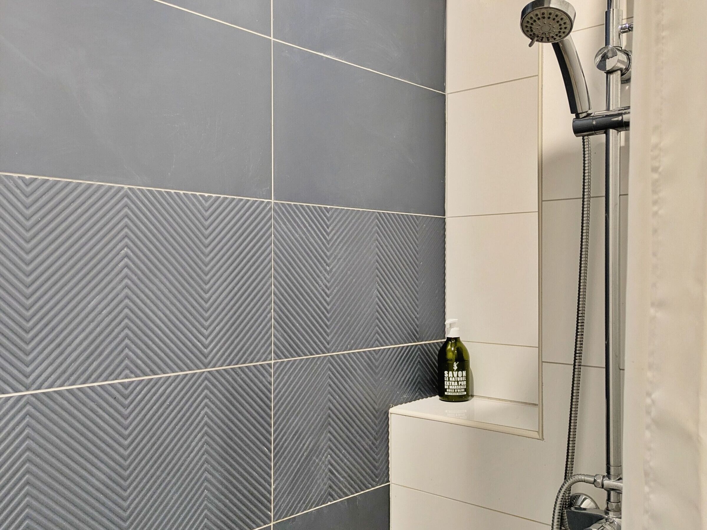 Een badkamer met een douche, een Savon flesje op een plank en een wand betegeld met een herringbone patroon.