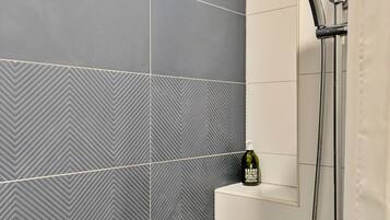 Een badkamer met een douche, een Savon flesje op een plank en een wand betegeld met een herringbone patroon.