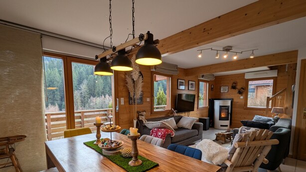 A cozy cabin interior with a wooden dining table, a sofa, a fireplace, and large windows offering a view of the forest. - Cozy chalet in Gérardmer with spa and sauna (Gérardmer)
