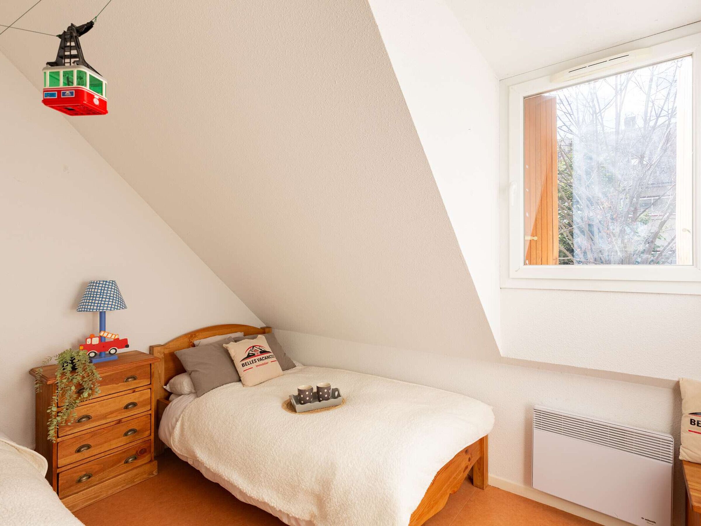 A bedroom with a sloped ceiling, a window with a view of trees, a bed with a white blanket, a wooden dresser, a lamp, and a small toy train.