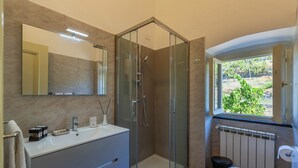 A modern bathroom with a glass-enclosed shower, a large mirror, a sink with toiletries, and a view of greenery through a window. - Cava Ardè Rooms:Garden view room, in the tranquility of Santa Giulia (Lavagna)