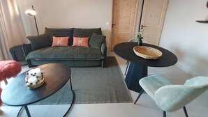 Una sala de estar acogedora con un sofá verde oscuro, dos almohadones, una mesita redonda con un objeto decorativo, un sillón de color claro y una puerta de madera grande al fondo.