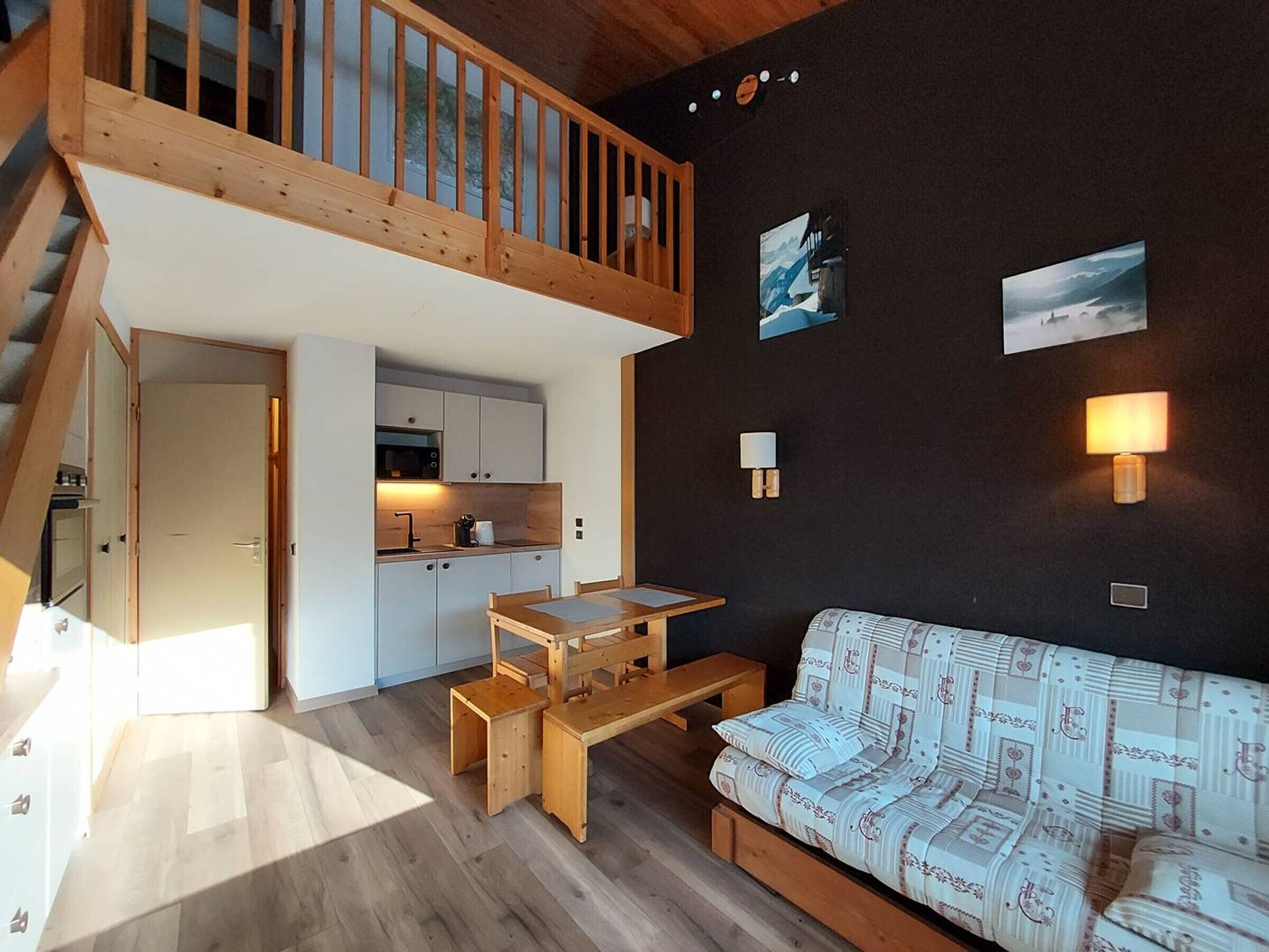 A compact living space with a wooden staircase, a kitchenette, a sofa, and wall-mounted lighting.