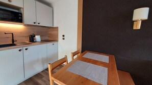 Pieni, käytännöllinen keittiö, jossa on valkoiset kaapit, musta mikroaaltouuni ja puinen pöytä sekä kaksi tuolia.