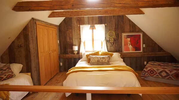 Une chambre mansardée chaleureuse avec un plafond en bois, un grand lit recouvert d’une couverture jaune, une armoire en bois et un tableau abstrait encadré.