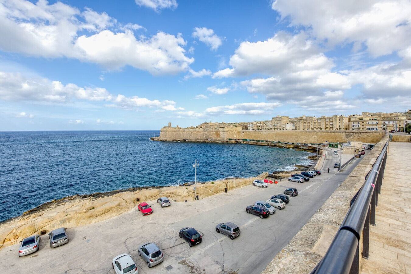 Una carretera costera con coches aparcados, un muro de piedra y vistas al mar y edificios al fondo.
