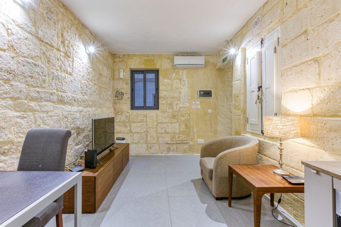 A compact living space with stone walls, a television on a wooden cabinet, a sofa, a table with a lamp, and a small window.