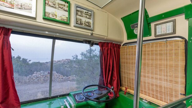 Interior d'un autobús amb volant, quadre de comandament i finestra amb cortines vermelles.
