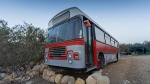 Un autobús immòbil estacionat en un camí de terra amb un límit rocos, envoltat de vegetació escassa i un cel serè.