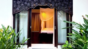 A room with a bed, a window with curtains, and a decorative wooden frame above the door. - Patmase Villas in Jimbaran (Badung)
