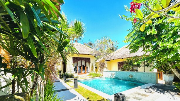 A pool area with a thatched-roof building, surrounded by lush greenery and a clear blue sky. - Patmase Villas in Jimbaran (Badung)