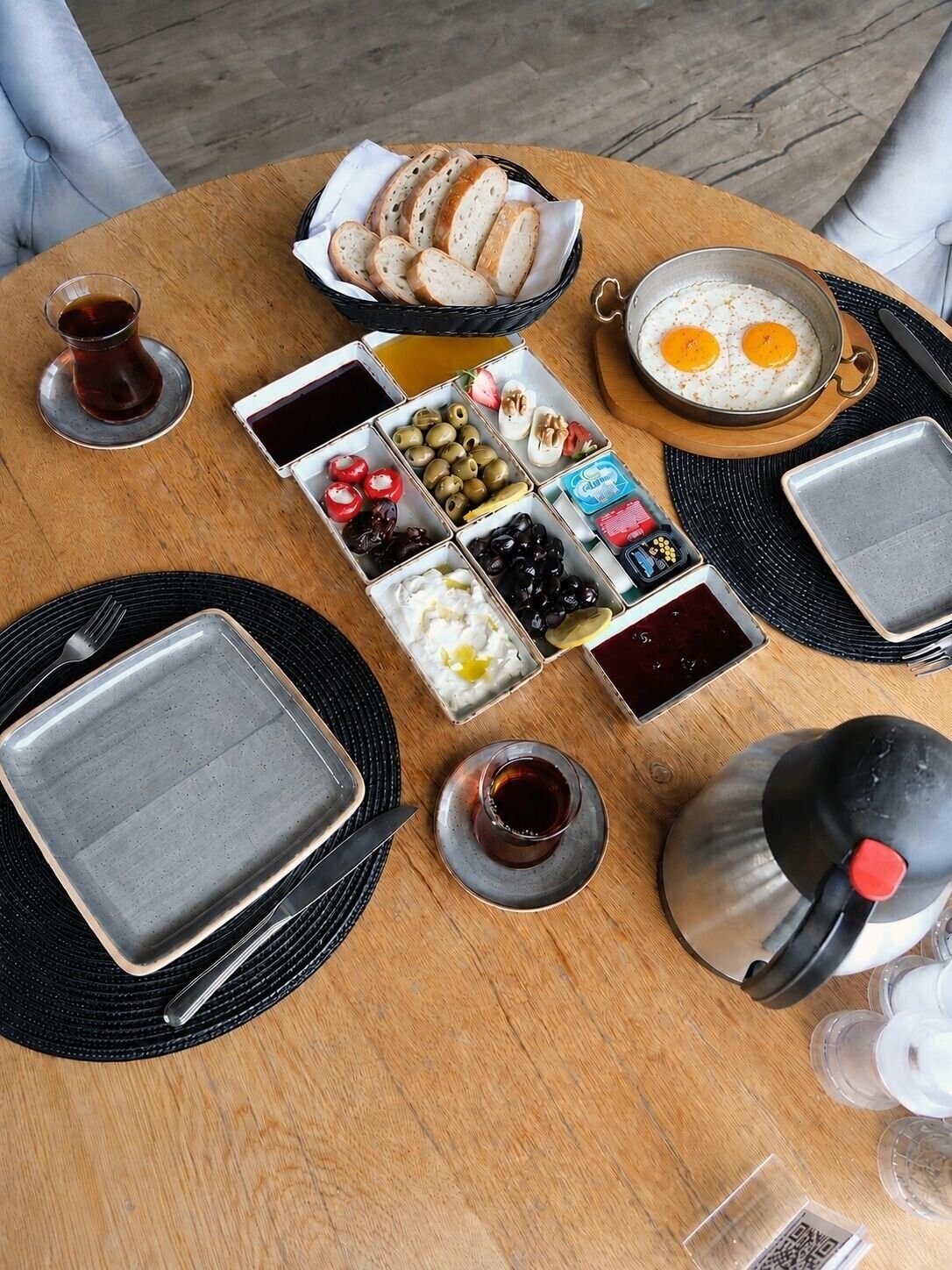 A breakfast setup with bread, eggs, olives, yogurt, and a coffee pot on a wooden table.