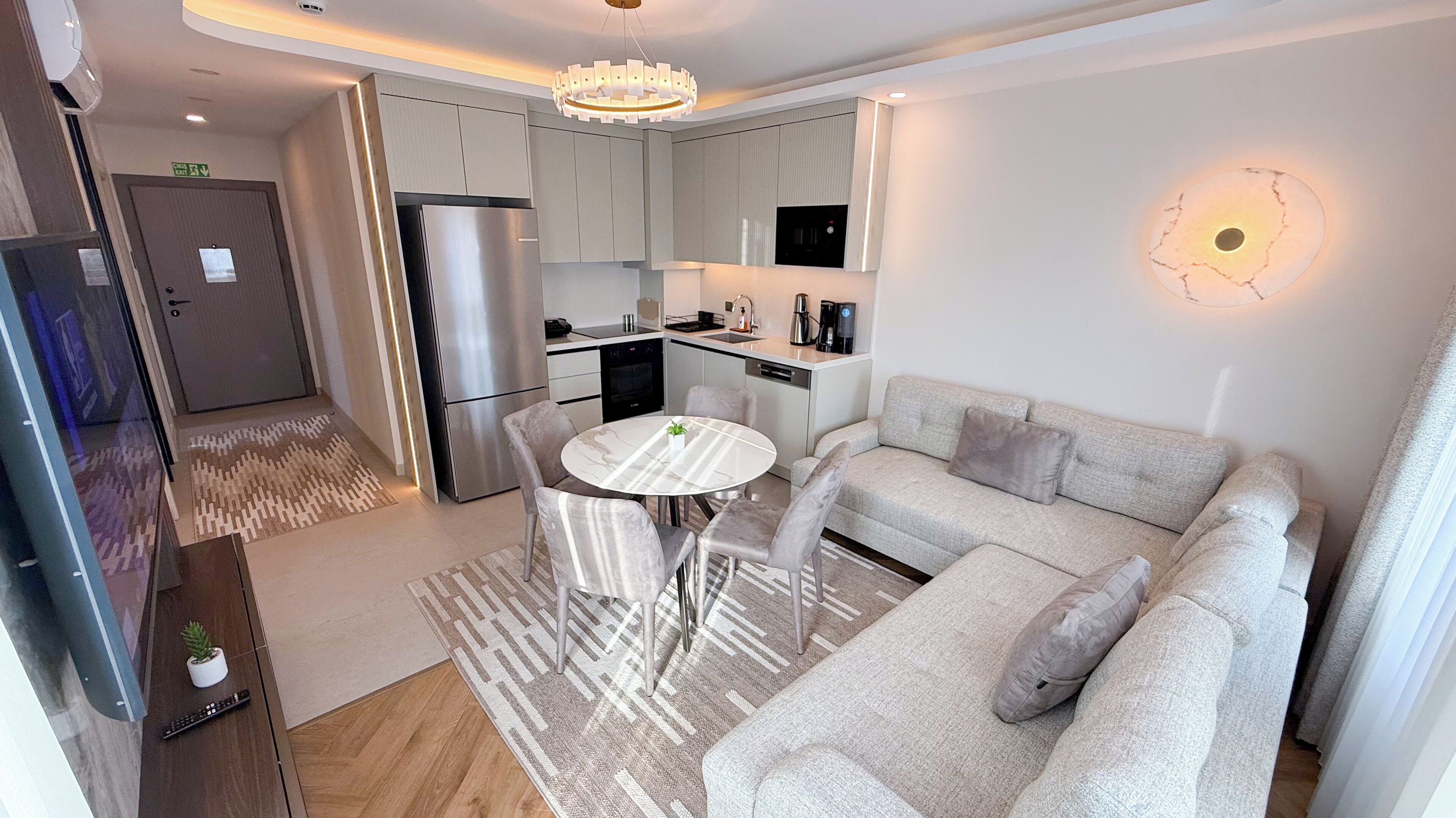 A modern kitchen with a built-in oven, a refrigerator, and a coffee machine. There is a dining area with a round table and chairs.