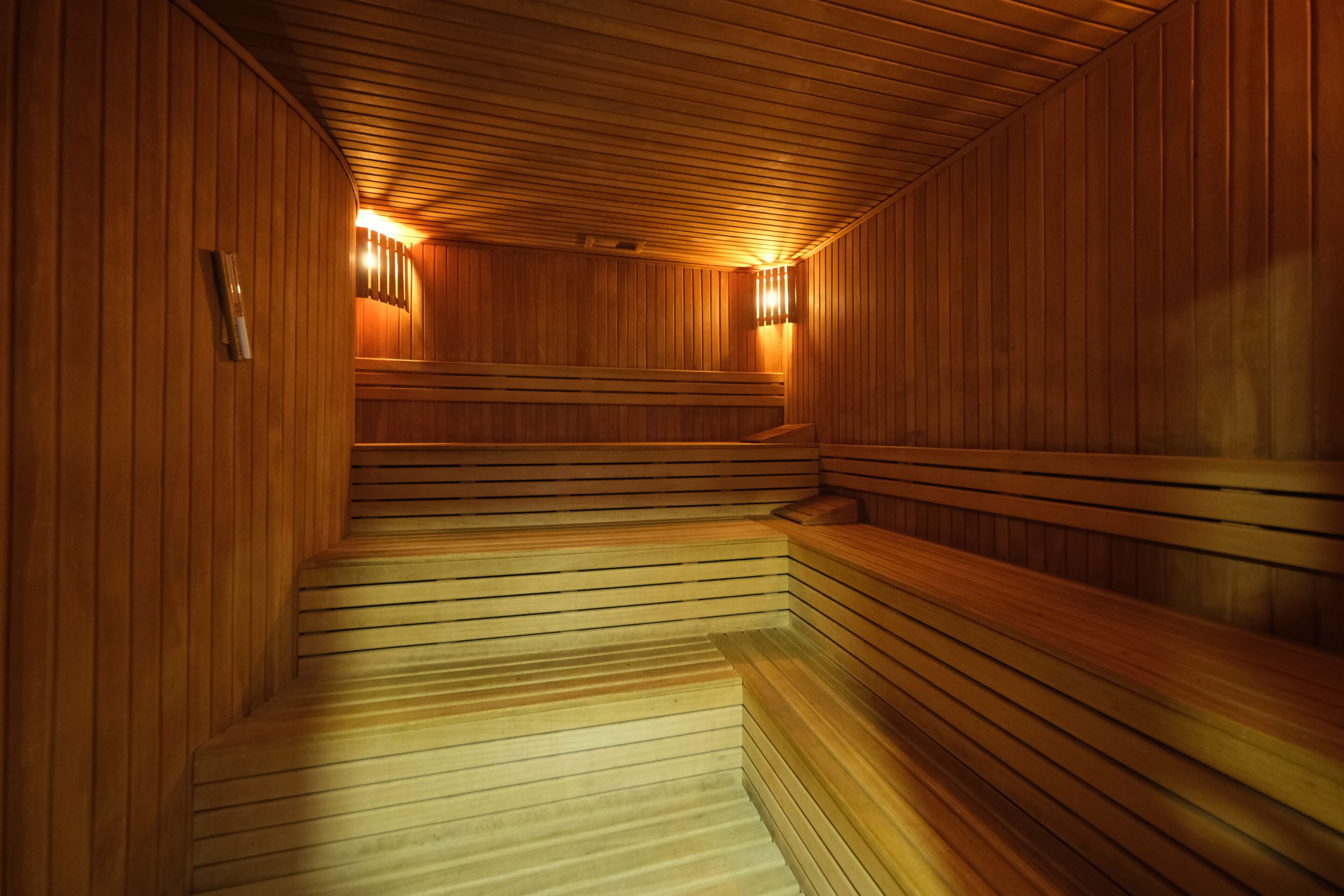 A sauna with wooden benches and sloped floors, wooden walls, and wall-mounted lights.