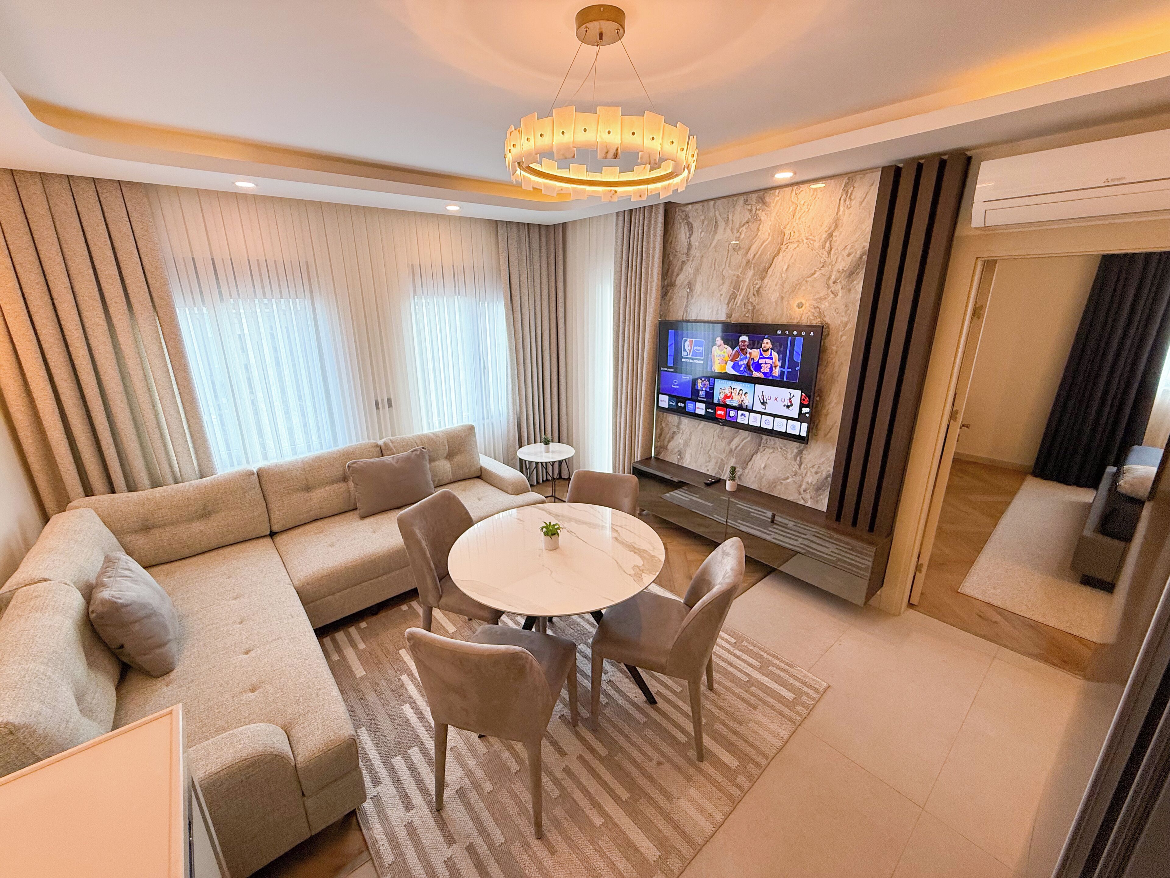 A modern living room with a beige sofa, a round dining table, and a TV mounted on the wall.