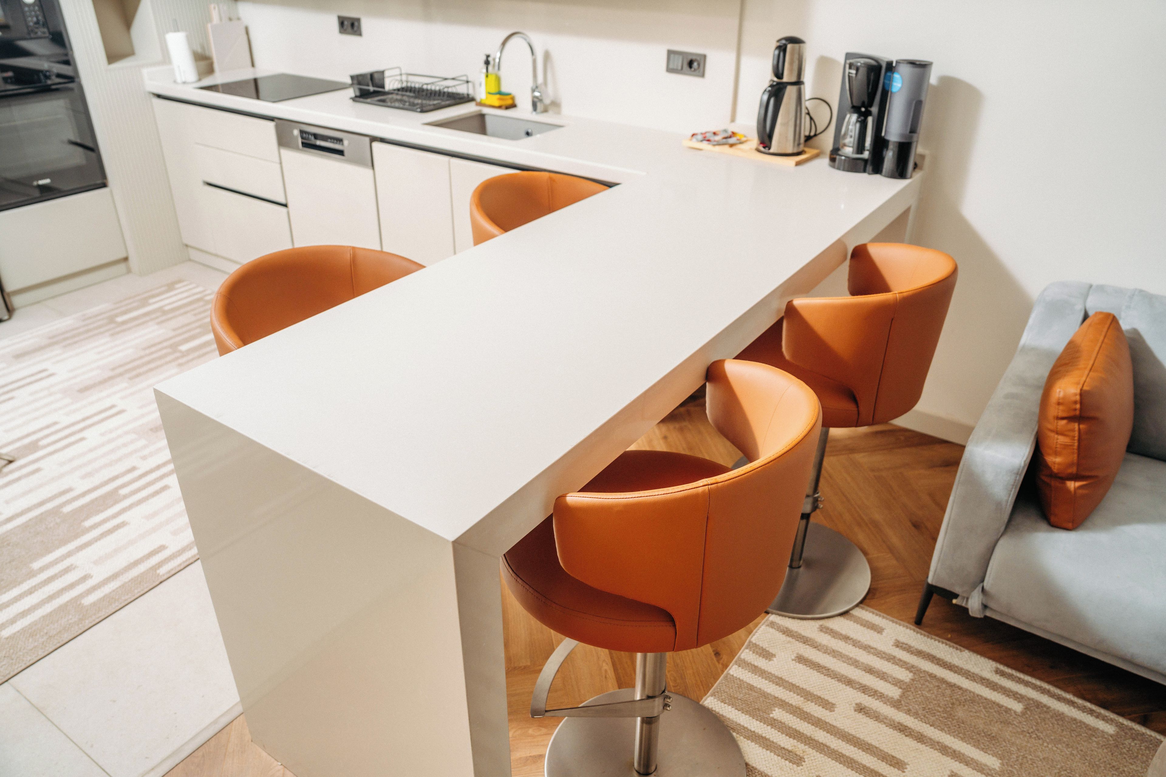 A modern kitchen with a white countertop, a built-in dishwasher, and a coffee machine. There are two orange chairs and a grey sofa with a cushion.